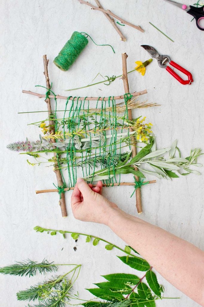 Hand weaving in a cutting of green leaves on almost completed nature weave made from twigs and green string.
