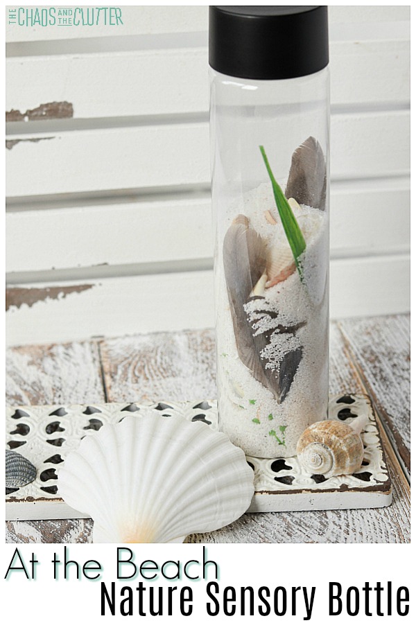 a bottle half filled with white sand, seashells, green seaweed, and a seagull feather sits on a distressed paint table with a few larger seashells. The text reads 