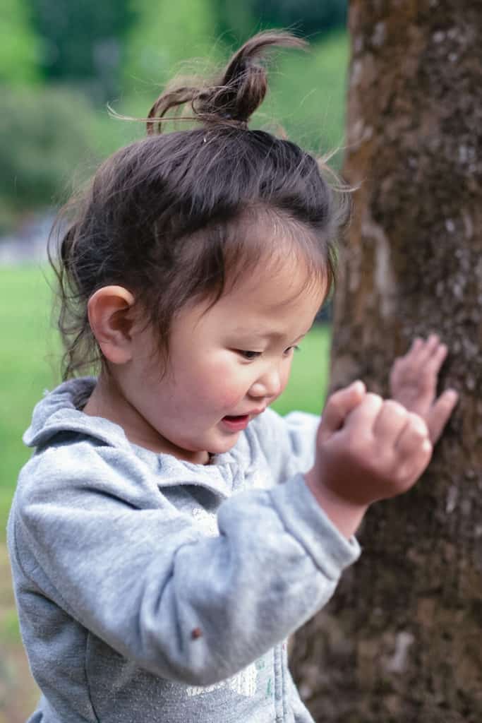 An 18-month-old toddler smiles while they touch the bark of a tree during a scavenger hunt.