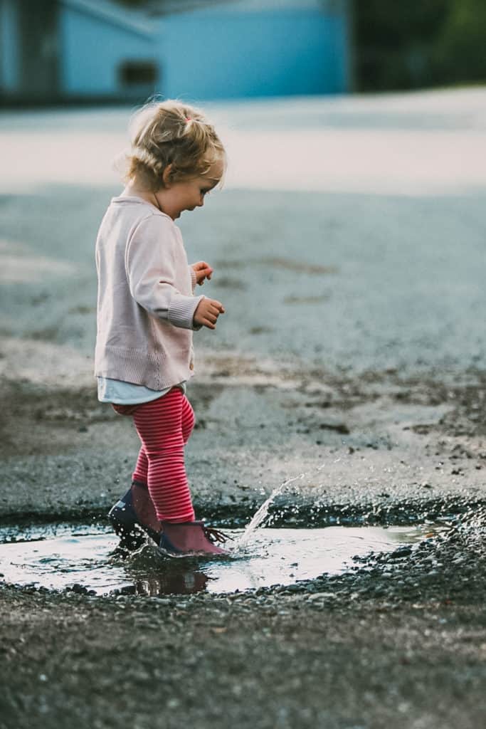 An 18-Month-Old toddler splashing water in a puddle while doing an outdoor learning activity.