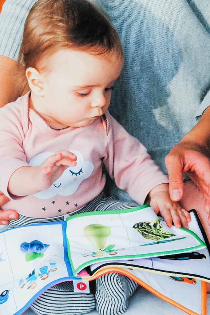 A baby wearing a nature shirt and looking at a nature book while sitting in a parent's lap.