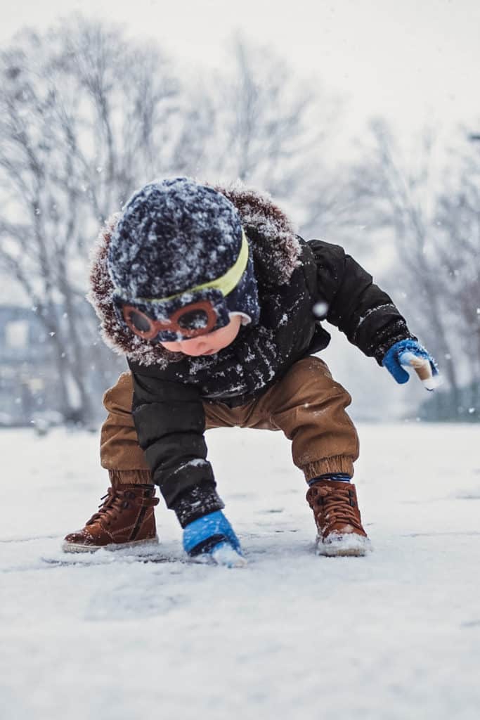 An 18-month-old toddler squats and leans over to touch snow with their gloved hand during an outdoor learning activity.