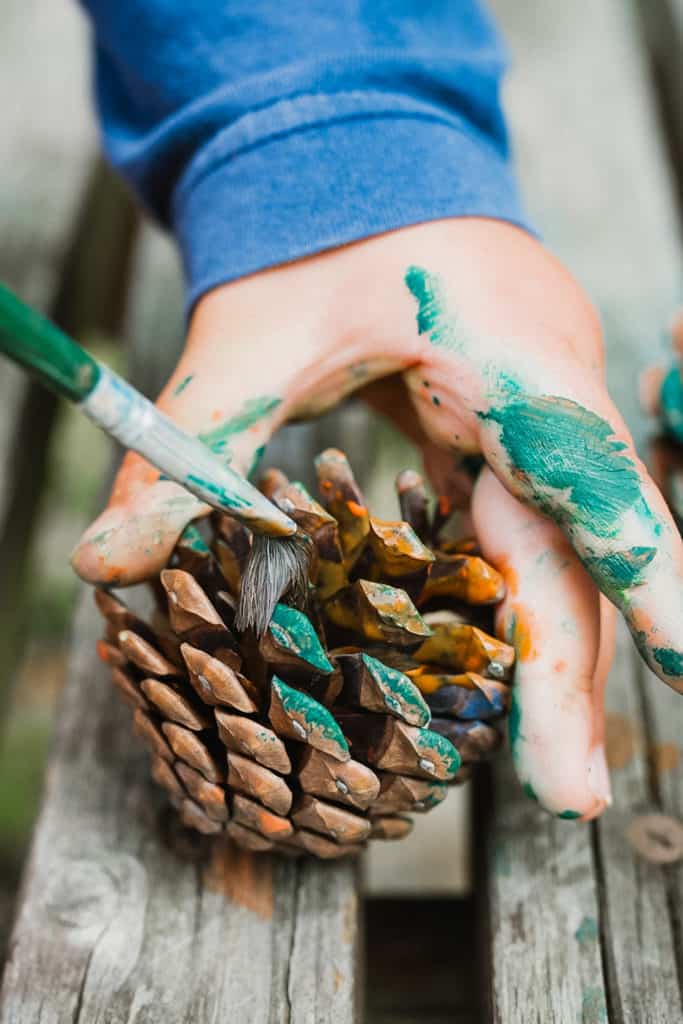 A toddler's paint-covered hand holds a paint-covered pinecone. A paintbrush adds more paint to the pinecone during an outdoor learning activity.