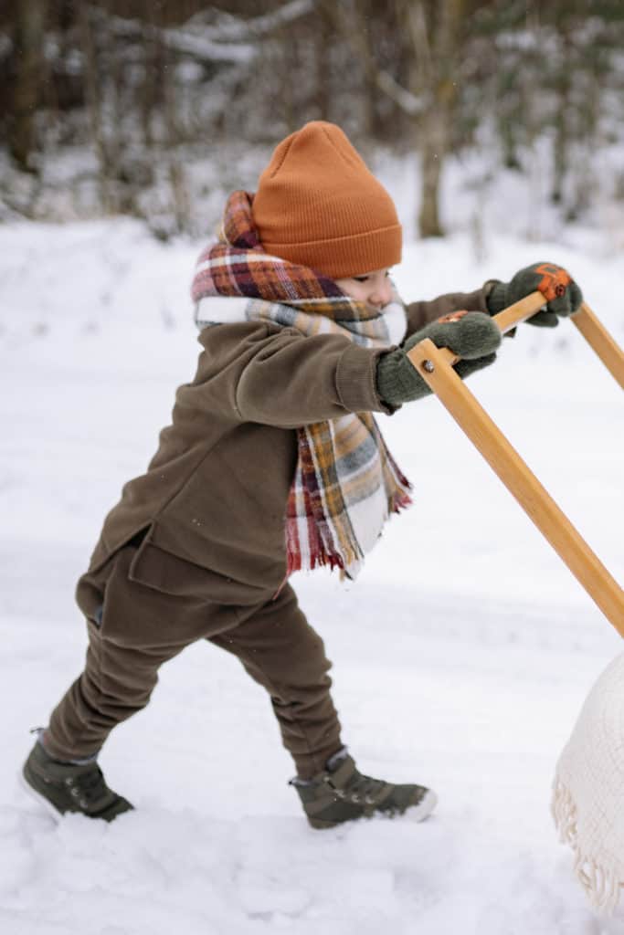 An 18-Month-old toddler pushing a stroller in the snow while developing physical skills during an outdoor learning activity.