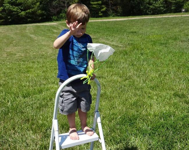 Child standing on a stepladder, dropping a toy attached to a paper parachute